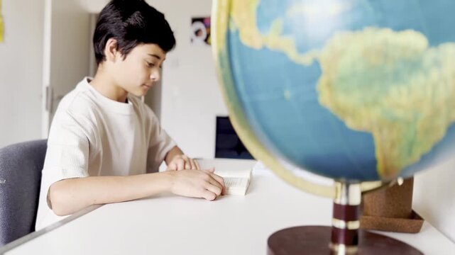 A school-age boy sits at a desk in the children's room and flips through the pages of a book. A boy with a book at a table with a globe.