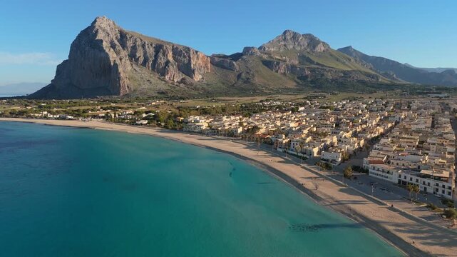 Clear blue water washes sandy beach of San Vito Lo Capo, Sicily, Italy. Rocky mountain rises above San Vito Lo Capo, Italy. 4K aerial shot reveals coastal architecture in San Vito Lo Capo, Italy