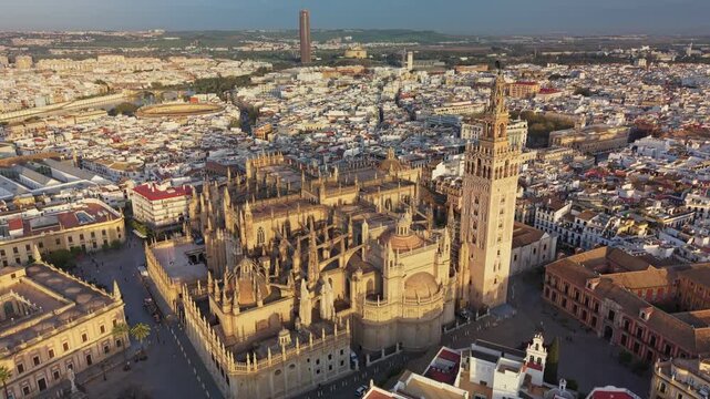 Giralda bell tower rising over Cathedral of Seville, Spain. Golden sunset hits Gothic roofs of cathedral in Seville, Spain. Dense cityscape surrounds historic cathedral in Seville, Spain. Aerial 4K