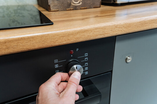 Hand Turning Oven Knob In Kitchen, Closeup Showing Fingers Gripping Chrome Dial, Wood Countertop Edge, Black Control Panel With Indicator Lights, Ambient Daylight, Home Cook Preparing Meal
