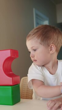 A boy is sitting at a table with colorful building blocks. The red and green blocks are stacked while the boy observes them with focus.