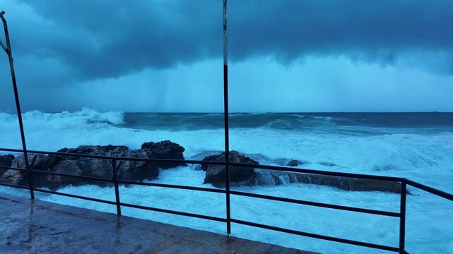 Stormy Mediterranean Coast Framed Through Pier Railing and Central Pole &ndash; Rocky Left Shore vs Open Turbulent Sea