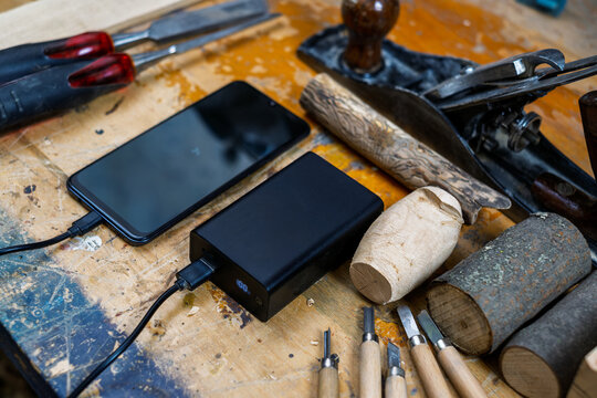 A wide high-angle shot of a phone connected to a 100% charged power bank, surrounded by chisels, wood planes, and logs.