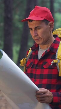 A man in a red cap examines a large map while standing in a forest. The man appears focused as he analyzes the map, surrounded by trees and nature.