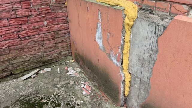 Cracked plaster corner sealed with foam, brick backdrop, vertical leak stain, damp mossy ground, scattered debris, construction