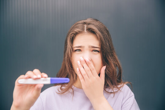 Young stylish trendy woman isolated over grey blue background. Shocked girl unexpected positive pregnance test result. Depression and anxiety. Looking straight on camera and showing test result.