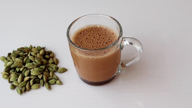 Close-up of a glass mug filled with hot milk tea next to a pile of green cardamom pods. This scene highlights traditional masala chai preparation and aromatic Indian spices on a white surface.
