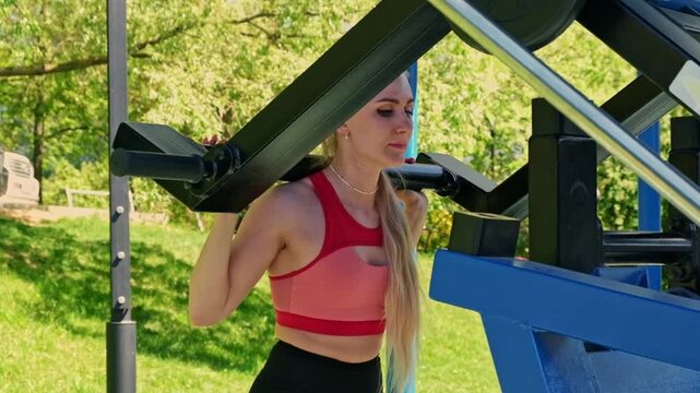 A bright side view captures a woman in sportswear doing a squat on public fitness equipment, surrounded by trees and grass. It supports active lifestyle, outdoor training, confidence, and strength.
