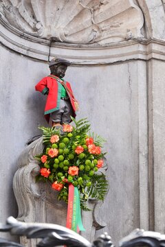 The "Manneken Pis" sculpture is dressed in vibrant red and green historical costume and adorned with a lush floral arrangement. This Brussels landmark is housed in a Rococo-style stone niche.