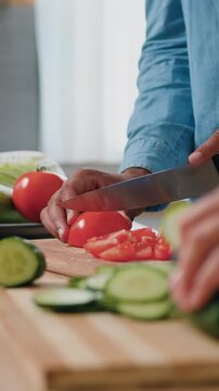 Close up shot. Male hands slicing a tomatoes and female hands cutting cucumber on a wooden cutting board for salad on a table with healthy food in the kitchen. Side view. Slow motion.