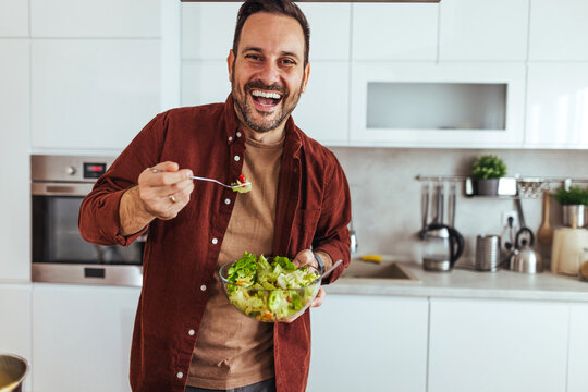 Happy Man Eating Fresh Salad in Modern Kitchen Holding Bowl and Fork, Smiling at Camera