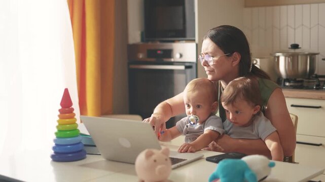 mother works remotely from home with two babies in her arms. pandemic remote work concept. mom tries to work at home in a business fun kitchen, small children interfere with sitting in her arms