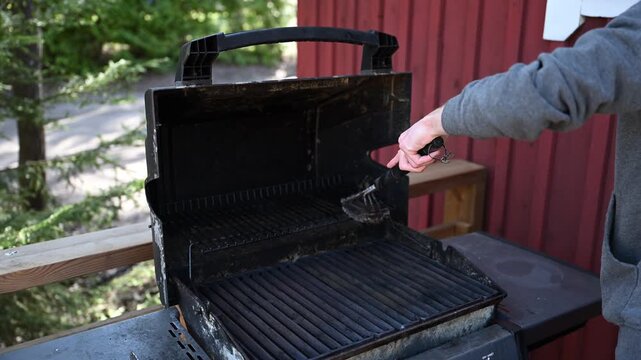 Cleaning outdoor barbecue grill with wire brush on wooden terrace in summer cottage backyard