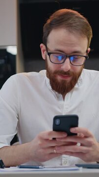 Cheerful business man sitting on desk and using a smartphone smiling in a modern kitchen. Freelancer male holding phone texting message technology app, typing email at home. Slow motion.