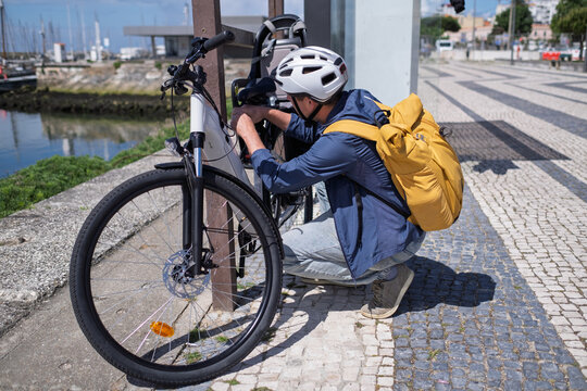 Young man in helmet securing his electric bicycle with a lock at a city bike rack near the water