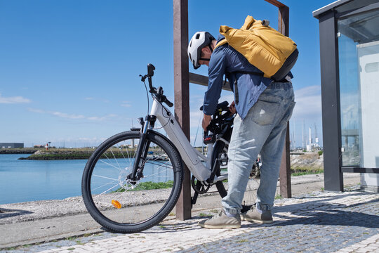 Young man in helmet securing his electric bicycle with a lock at a city bike rack near the water