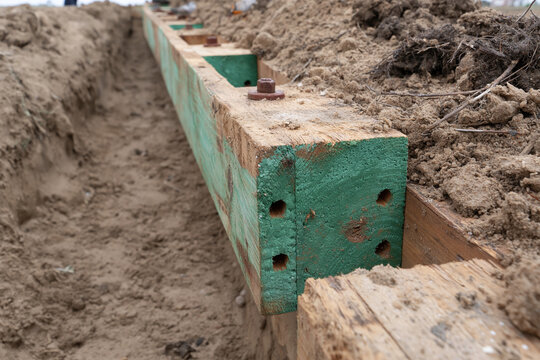 Green-painted wooden formwork stands in dirt. Holes and fasteners show construction effort. Earth surrounds the structure in an open trench. This image captures early building groundwork