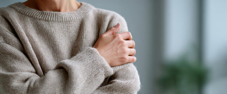 Close-up of person wearing cozy beige knitted sweater holding their shoulder indoors with blurred background