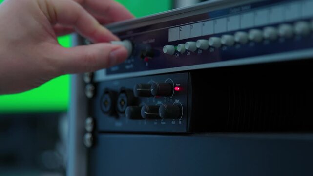 A sound engineer pushing buttons on a mixer to control it. A black keyboard sits on a white desk next to a black box with a green background.