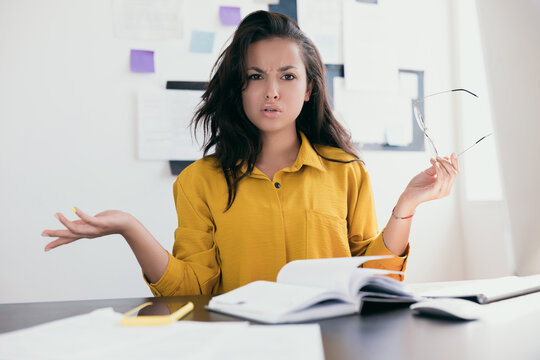 Perturbed office worker. Stressed young woman throw up her hands. Lady in yellow blouse holding glasses in hand. There is an open notebook and phone on the table. Office work work from home concept.