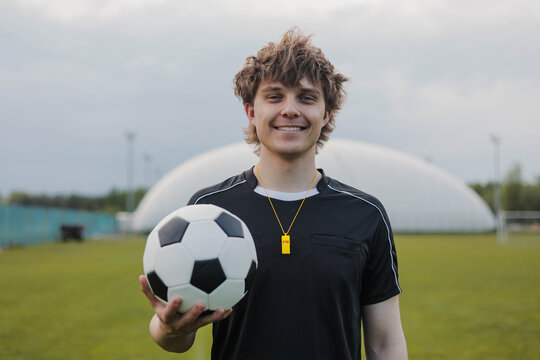 Young smiling soccer referee or coach holding a football in black referee uniform. Concept of officiating, fair play, sports rules and game control.