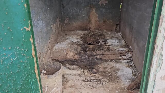 Old dirty outdoor rural toilet with broken green wooden door and concrete floor hole in abandoned village yard showing poor sanitation conditions
