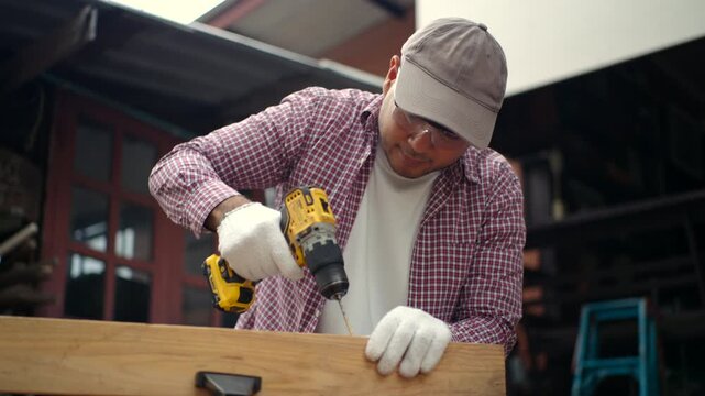 Confident carpenter man using cordless power drill to make hole in laminated wood board inside timber workshop. DIY woodworking, handmade furniture, home improvement and craftsmanship concept.