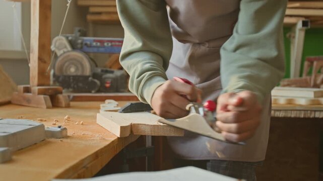 Midsection view of unrecognisable teenager wearing light green long-sleeved shirt and beige apron shaping wooden detail with hand plane while doing woodwork