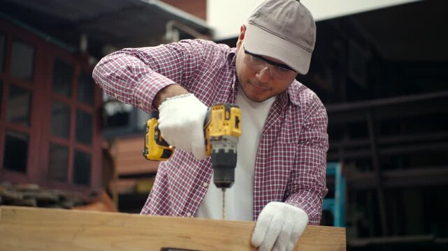 Confident carpenter man using cordless power drill to make hole in laminated wood board inside timber workshop. DIY woodworking, handmade furniture, home improvement and craftsmanship concept.