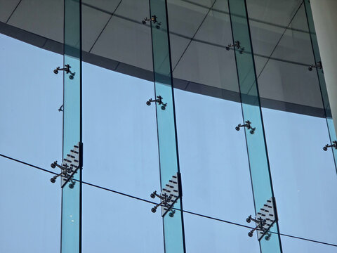 Modern glass facade of commercial building featuring structural spider fitting connection under calm clear sky reflecting imposing industrial minimalist architectural exterior design details