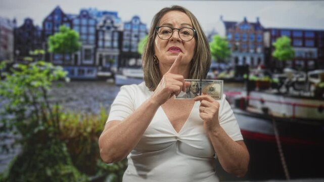 Elderly woman wearing glasses holds us dollar banknote and makes finger to lips shushing gesture in studio set; silence.