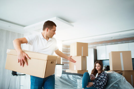 Moving and relationship concept. He is not satisfied. The husband unpacks the boxes. And the wife meanwhile does nothing. She prefers to talk to her friends. The husband is not satisfied.