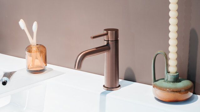 White countertop holds a modern sink. Copper faucet has a sleek handle. Two toothbrushes sit in a glass. Toothpaste tube rests beside them. Textured candle is in a ceramic holder