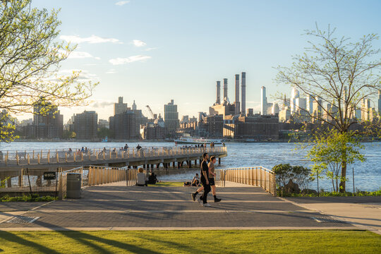 View of people walking along the Williamsburg waterfront park with the East River and Manhattan skyline in the background during golden hour Kings County, New York, United States.