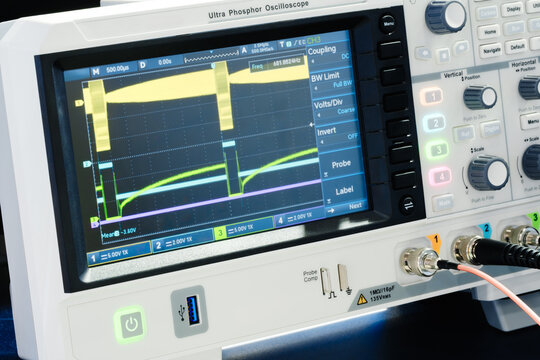 Digital oscilloscope displays multiple colored waveforms. Yellow and green lines show signal patterns on the screen. Blue and purple traces indicate electrical data