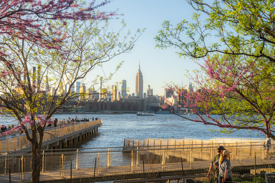 View of the Manhattan skyline with the Empire State Building framed by spring blossoms and people on a waterfront pier in Kings County, New York, United States.