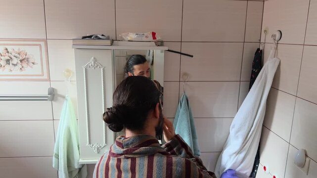 Adult bearded Caucasian man in a striped bathrobe leaning over the sink to rinse his mouth and wash his face during a quiet morning routine at home authentic slow life lifestyle reflection shot