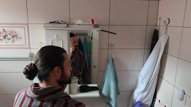 Adult bearded Caucasian man with long hair in a bun wearing a striped bathrobe brushing his teeth with a toothbrush in front of a mirror during his morning hygiene routine at home reflection shot