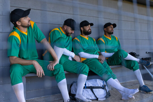Diverse male team sitting on bench in dugout wearing uniforms and caps near duffel bag