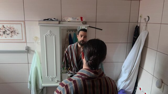 Bearded Caucasian man in a striped bathrobe inspecting his face in a bathroom mirror then starting to brush his teeth as part of a daily morning hygiene routine at home authentic lifestyle shot
