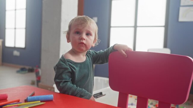 Little boy in green shirt rests hand on pink chair by red table with markers and crayons inside building; quiet curiosity.