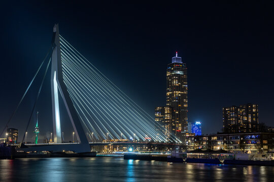 View of the illuminated Erasmusbrug and city skyline at night with the De Zalmhaven skyscraper reflecting on the water in Rotterdam, Zuid-Holland, Netherlands.