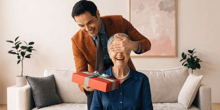 Joyful man surprises elderly woman with gift in cozy living room, creating warm and happy moment together