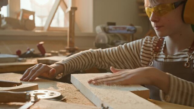 Blond teenage boy wearing yellow safety goggles, earmuffs and striped shirt blowing sawdust off wooden plank in woodworking school