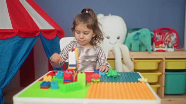 Girl stacks colorful toy blocks with her hands on a play table in a children's playroom inside a building; curiosity.