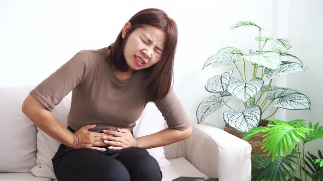 Asian woman sitting on a sofa suffering from severe stomach ache and abdominal pain. Concept of period cramps, food poisoning, gastritis, or digestive problems with a painful facial expression