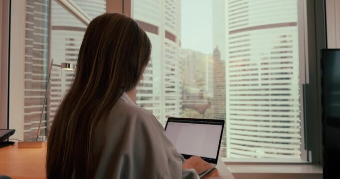 Woman typing on laptop at desk in modern high-rise office overlooking skyscrapers in Hong Kong during daytime. Over-the-shoulder view of remote work or corporate business. Slow motion.