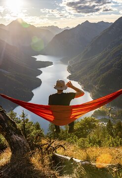 Man with a sun hat sitting in an orange hammock, panoramic view of mountains with lake, Plansee, Tyrol, Austria
