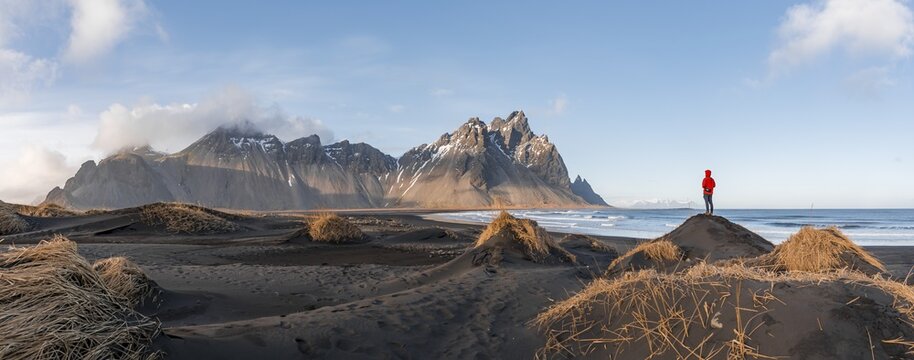 Man in red jacket standing on dunes with dry grass, black lava beach, sandy beach, mountains Klifatindur, Eystrahorn and Kambhorn, headland Stokksnes, mountain range Klifatindur, Austurland, East Iceland, Iceland