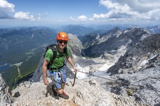 Mountaineer with helmet in a secured via ferrata, Zugspitze via ferrata, ascent to the Zugspitze, view of mountain basin with snowfield of the H&ouml;llentalferner and H&ouml;llental, Wetterstein range, Wetterstein range, Bavaria, Germany